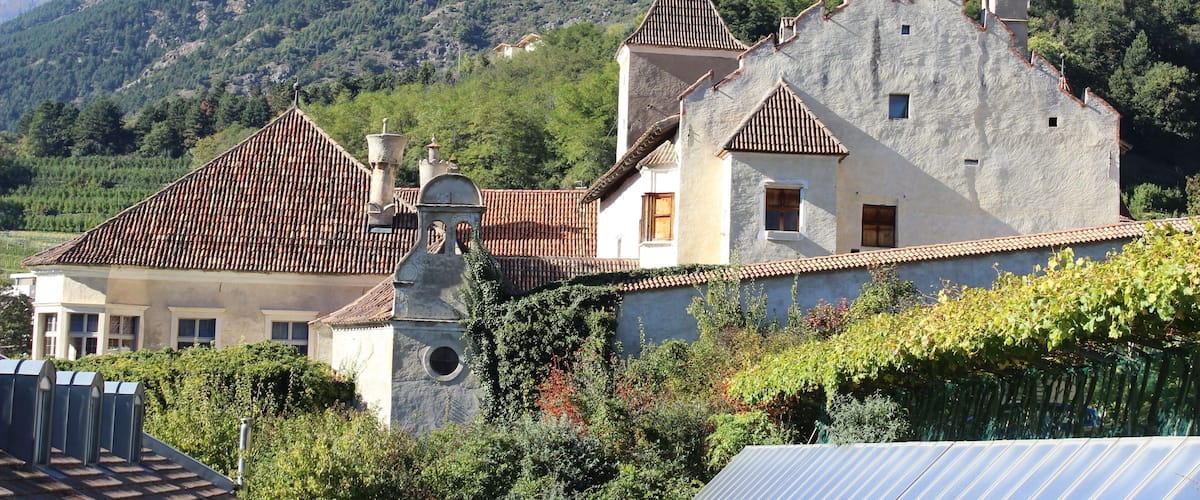 A traditional stone house with a tiled roof, surrounded by lush greenery and a mountainous backdrop.