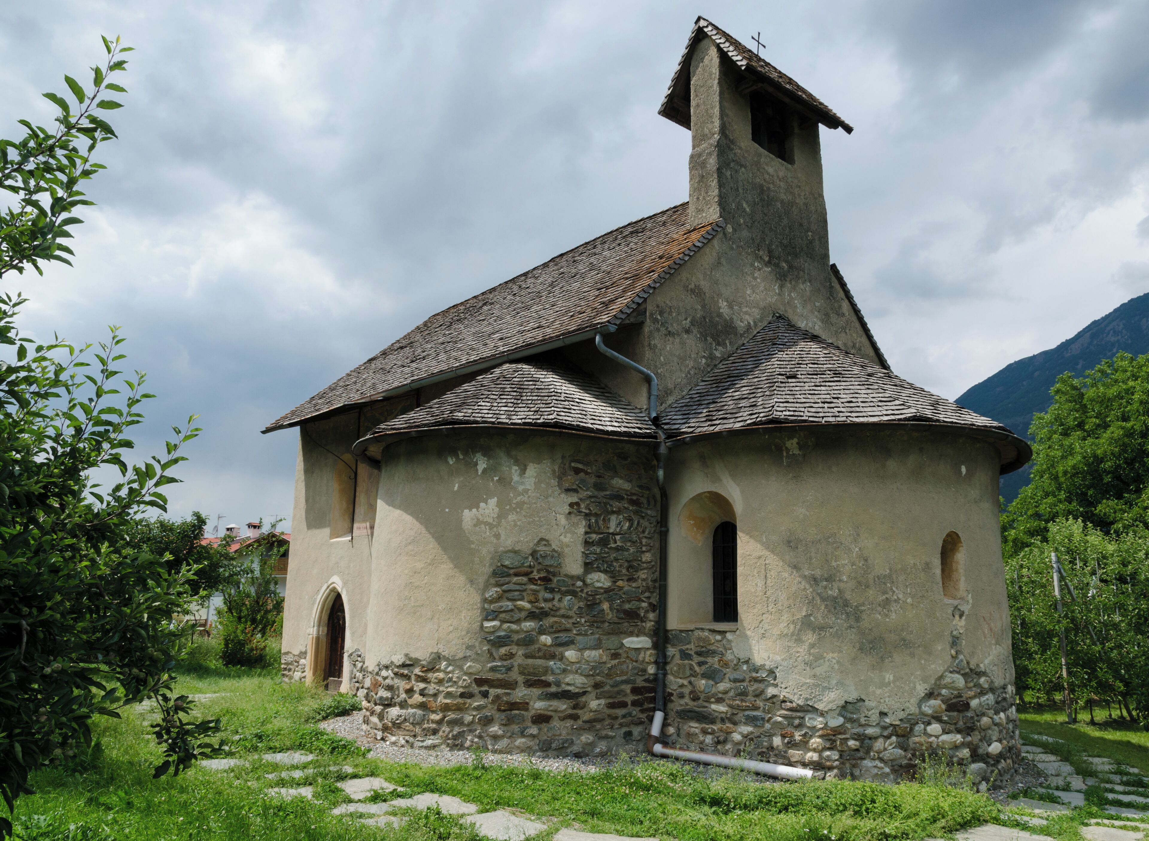 A small stone church with a steep roof and a cross on top, surrounded by greenery and mountains.