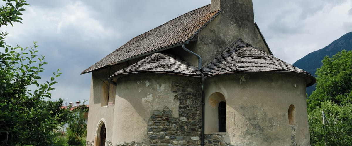A small stone church with a steep roof and a cross on top, surrounded by greenery and mountains.