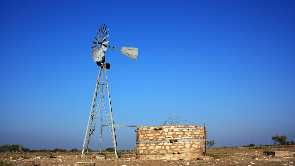 A windmill with a stone base and metal structure, set in a dry, rocky landscape with sparse vegetation.
