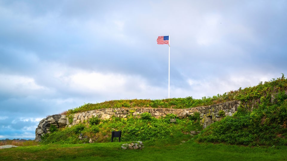 A stone wall with an American flag on a flagpole, surrounded by greenery and a cloudy sky.