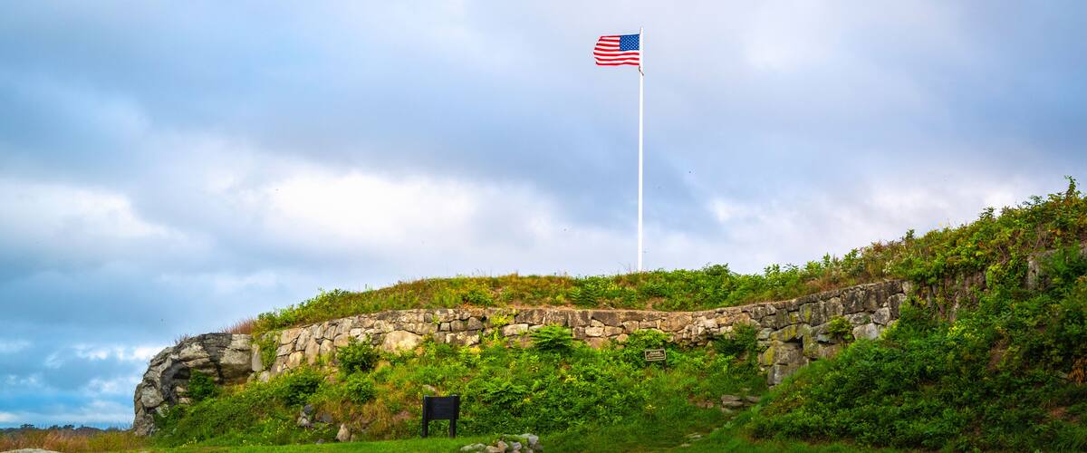 A stone wall with an American flag on a flagpole, surrounded by greenery and a cloudy sky.