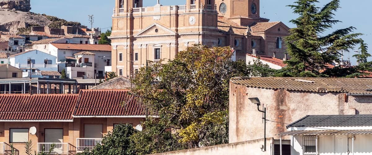 A historic church with two bell towers, surrounded by residential buildings and a hill in the background.