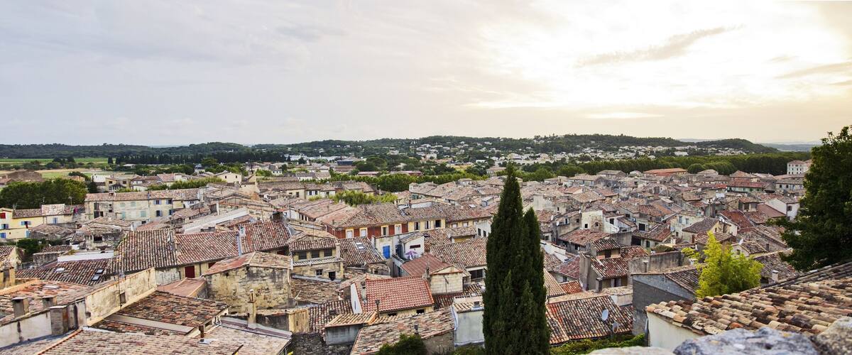 A densely packed historic town with terracotta-roofed buildings, a prominent tall cypress tree, and rolling hills in the background.