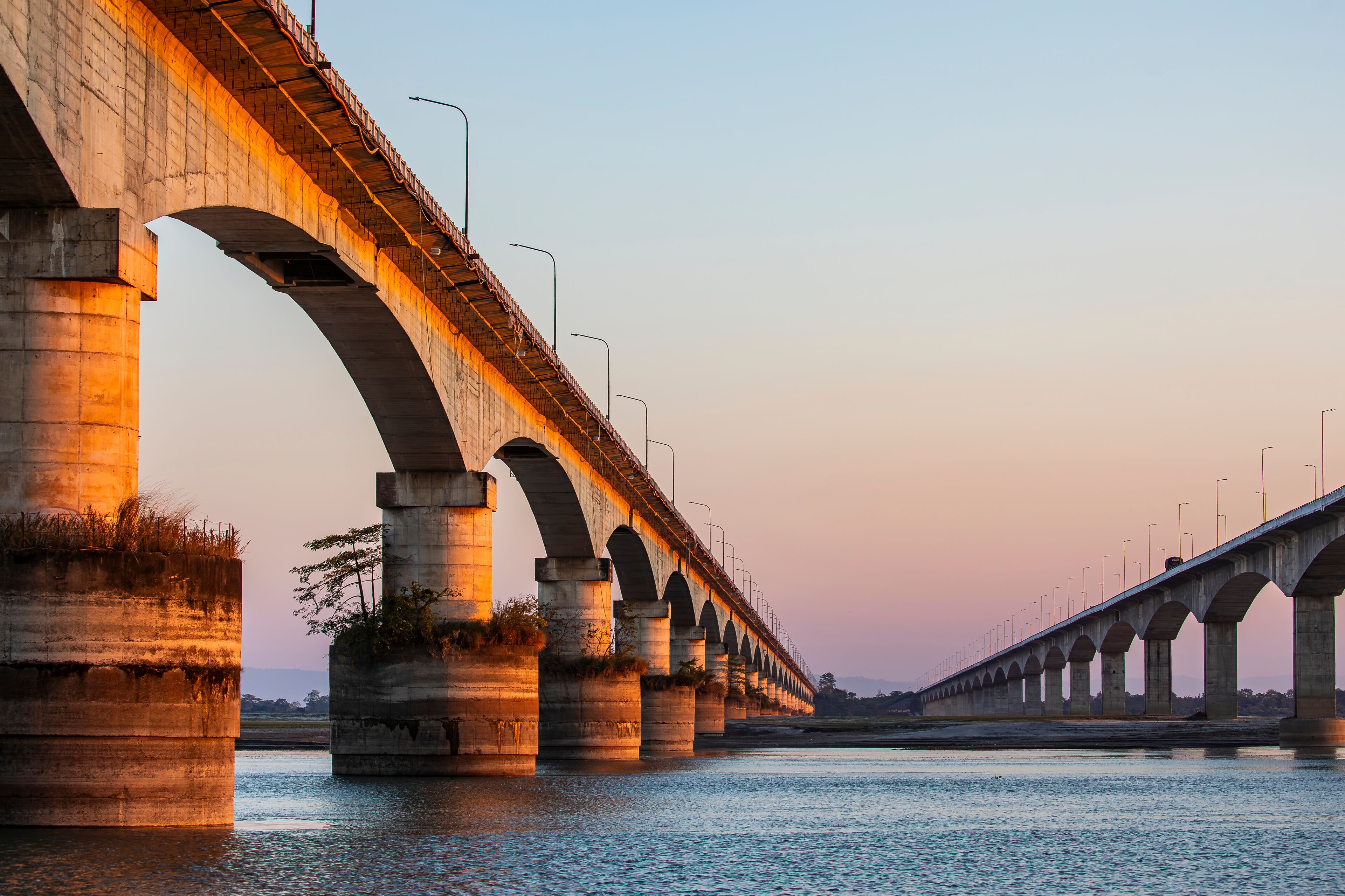 Sunset over the Brahmaputra River, with the Koliabhomora Bridge silhouetted against warm evening light and reflections shimmering on the water.