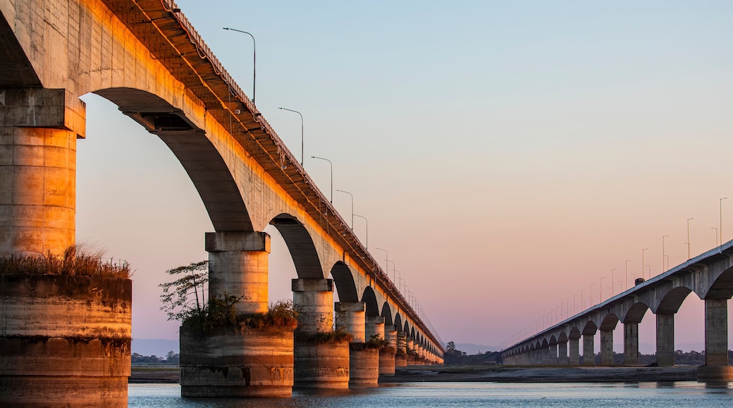 Sunset over the Brahmaputra River, with the Koliabhomora Bridge silhouetted against warm evening light and reflections shimmering on the water.