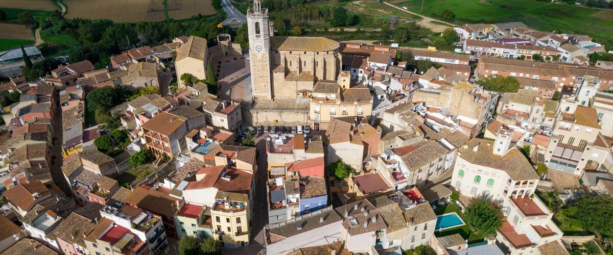 An aerial view of a historic town with a prominent church tower, surrounded by tightly packed buildings and green fields.