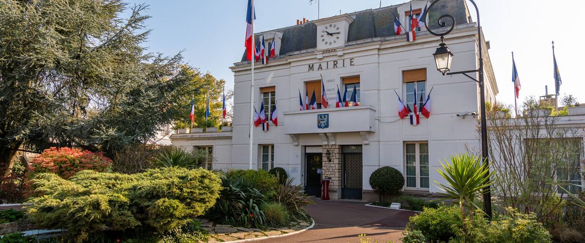 A building with 'MAIRIE' on the facade, surrounded by lush greenery and multiple French flags.