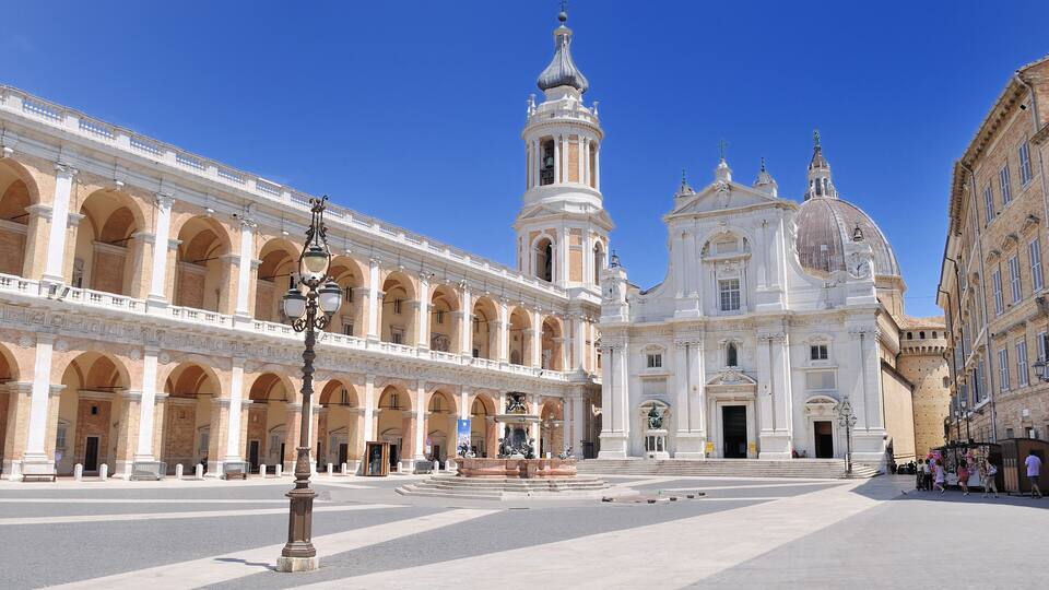 A large open plaza with a prominent white church with a tall bell tower and a fountain in the center.