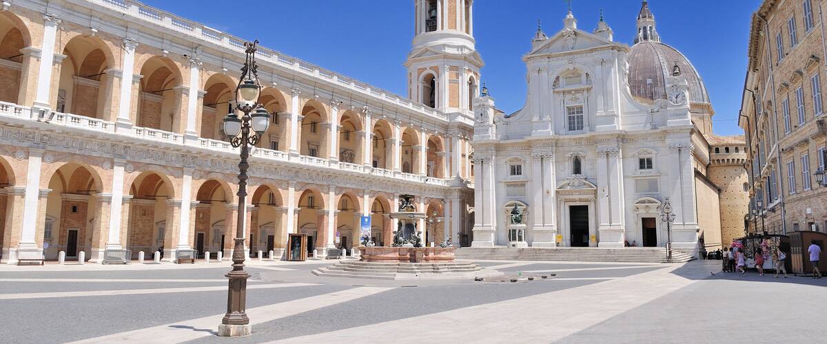 A large open plaza with a prominent white church with a tall bell tower and a fountain in the center.
