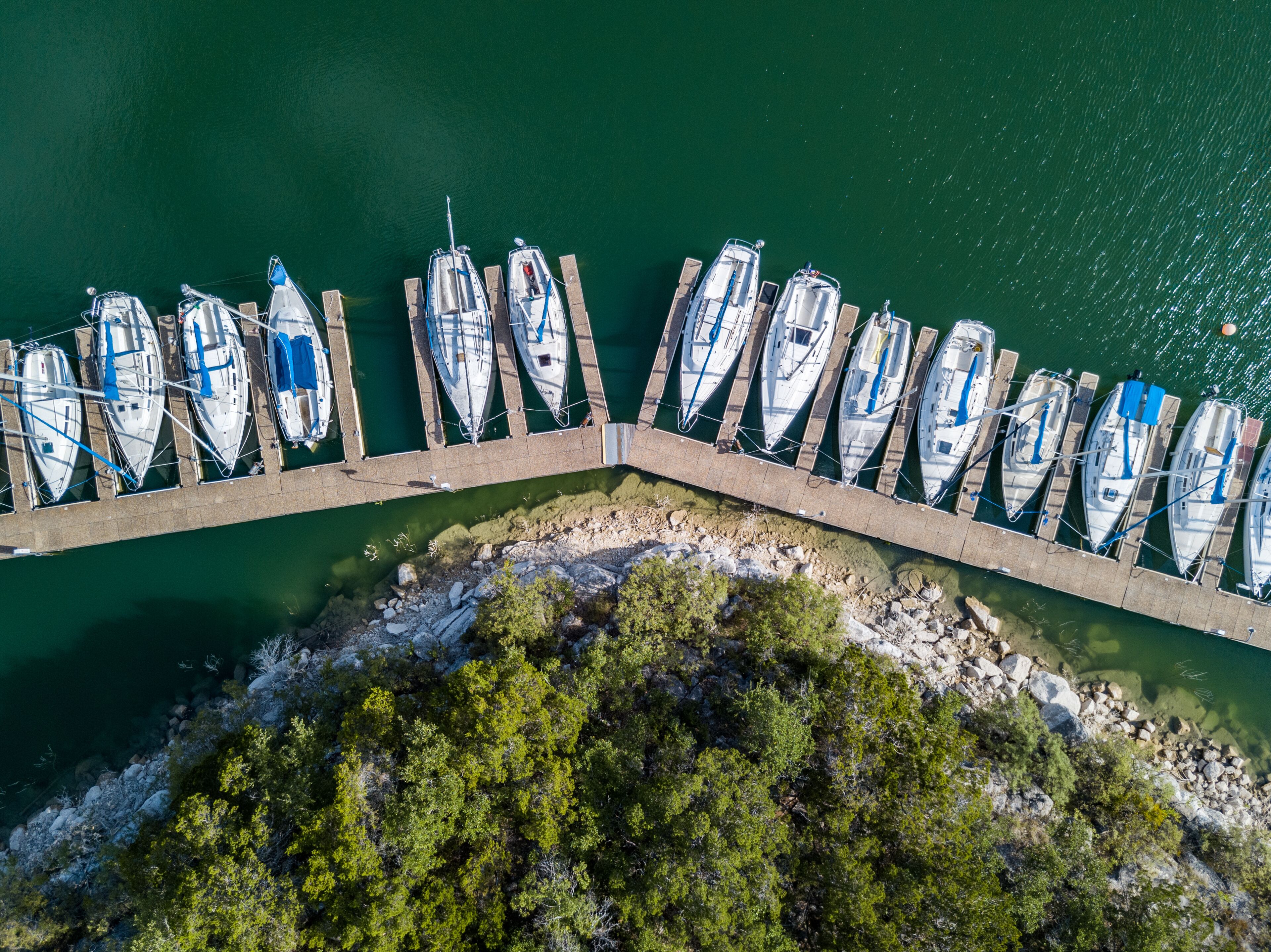 Un muelle con varias embarcaciones amarradas en fila, rodeado de agua tranquila y una costa rocosa con vegetación densa.