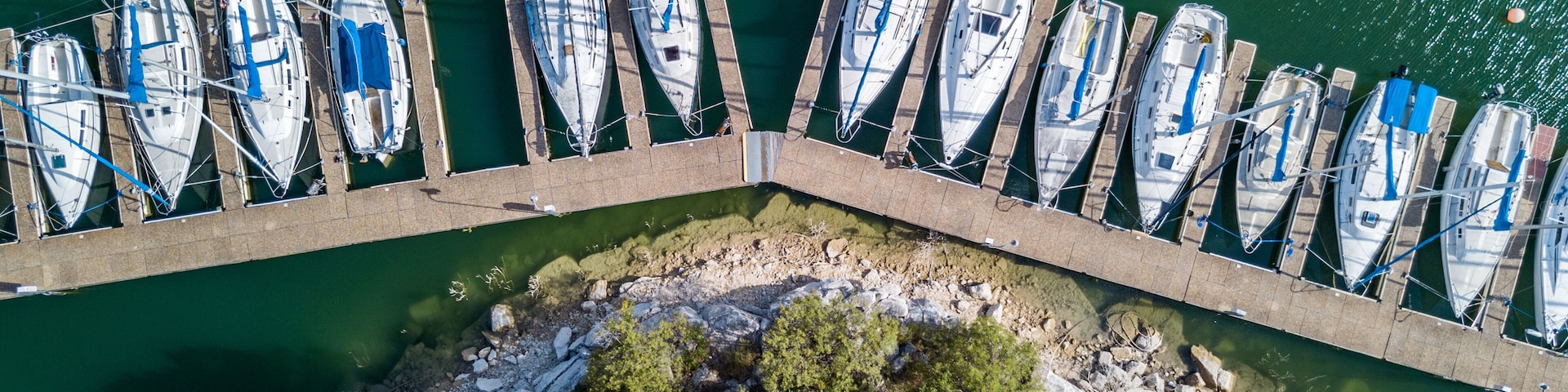 Un muelle con varias embarcaciones amarradas en fila, rodeado de agua tranquila y una costa rocosa con vegetación densa.