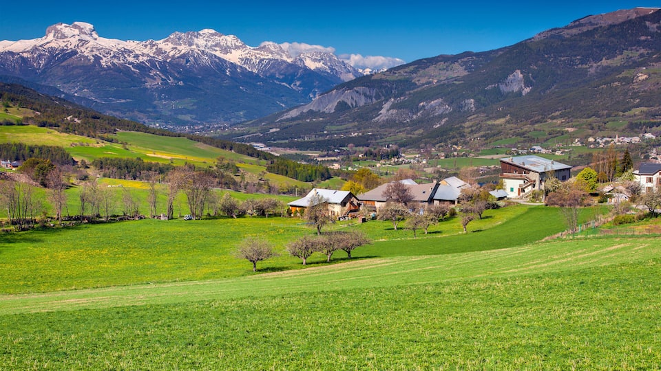 A mountainous landscape with a village nestled among green fields and snow-capped peaks in the background.