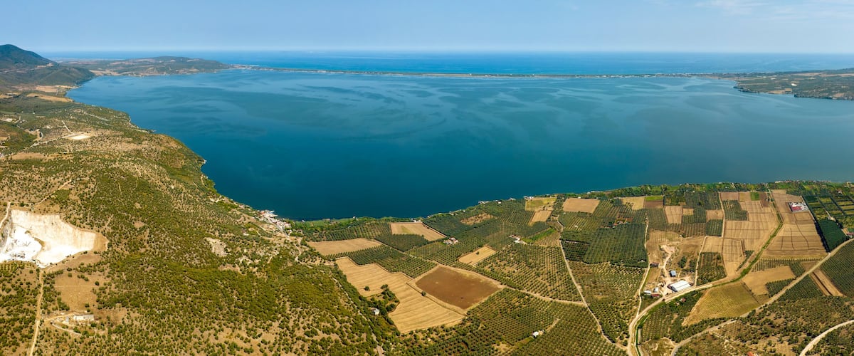 An aerial view of a coastal landscape with a large body of water, terraced fields, and a mountainous backdrop.