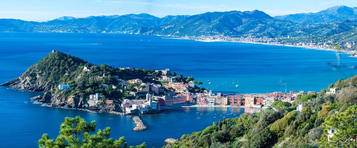 A coastal town with a prominent hilltop settlement, a clear blue sea, and distant mountains.