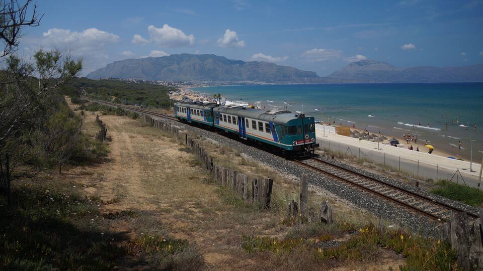 A train traveling along a track near a beach with mountains in the background.