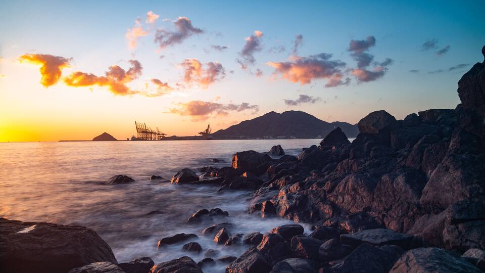 A rocky shoreline at sunset with a distant pier and mountains.