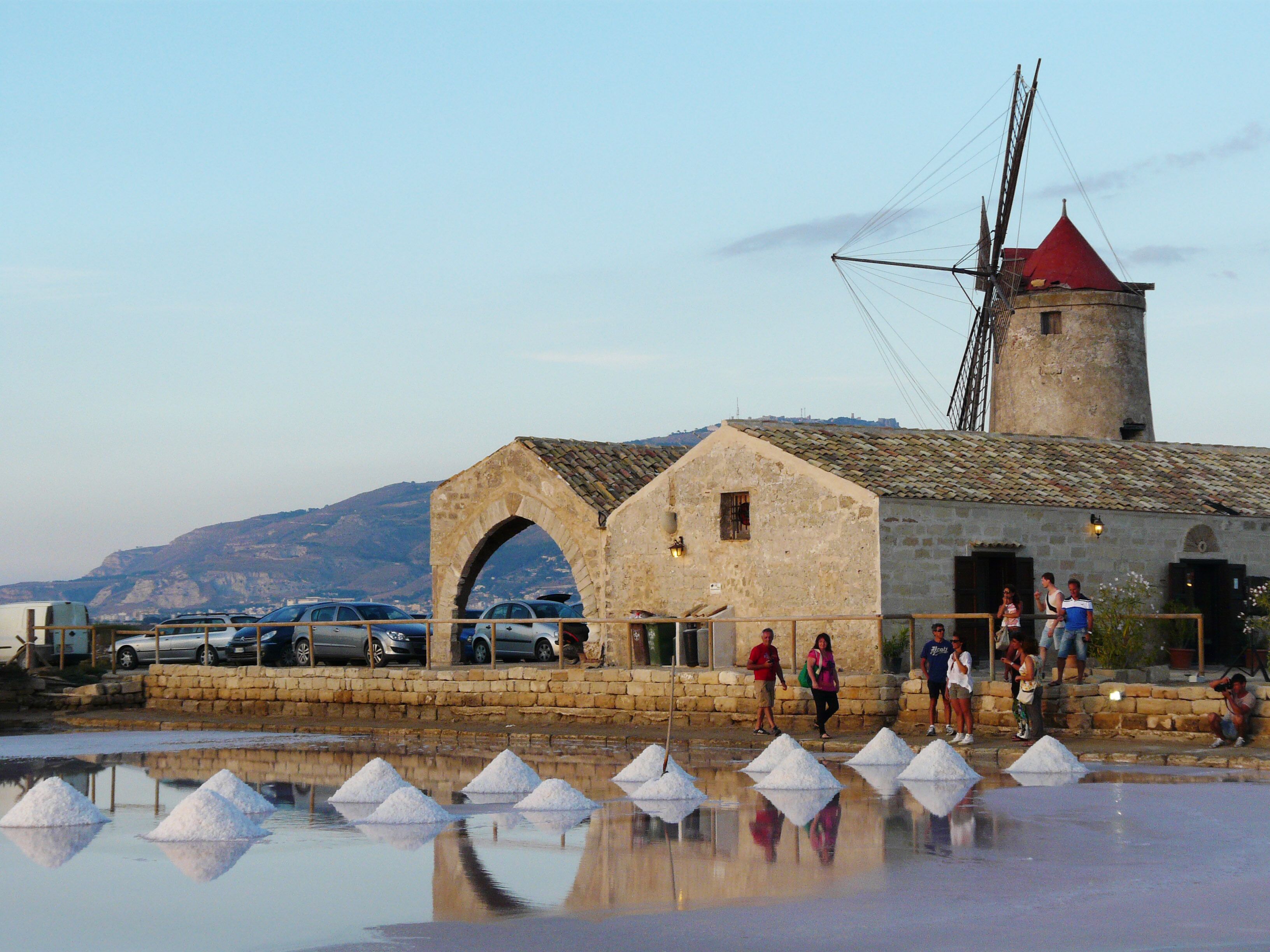 A historic building with a red roof and a windmill, a stone archway, and a salt evaporation pond with white salt piles.