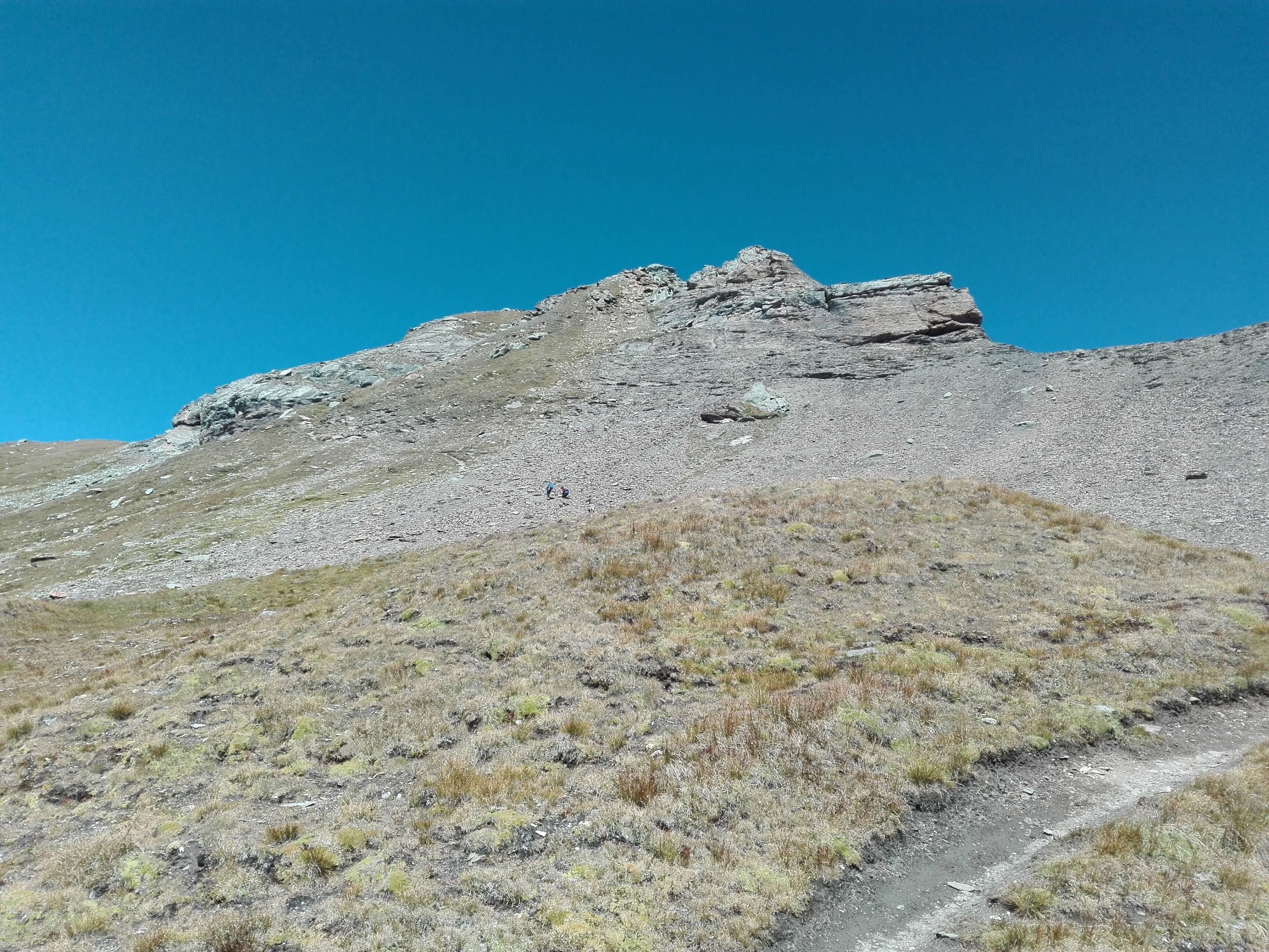 A rocky mountain slope with sparse grass and a clear blue sky.