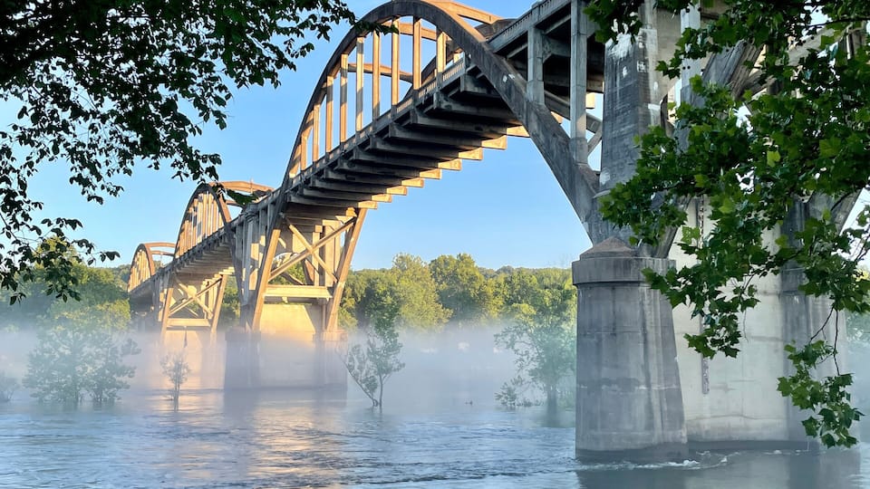 A steel bridge over a river with steam rising from the water, surrounded by greenery.