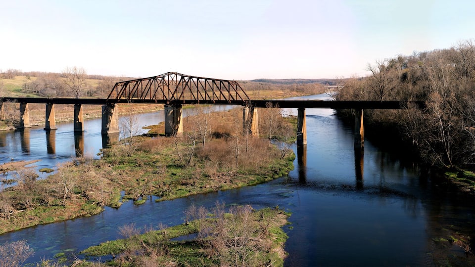 A steel bridge over a river with surrounding trees and hills.