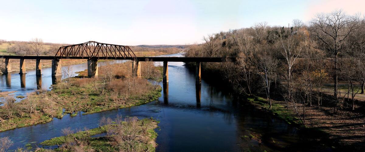 A steel bridge over a river with surrounding trees and hills.
