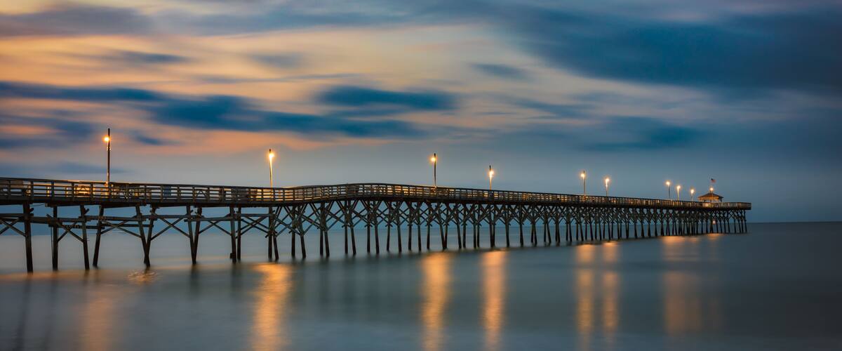 A long pier extending into calm waters at dusk with lights along its length.