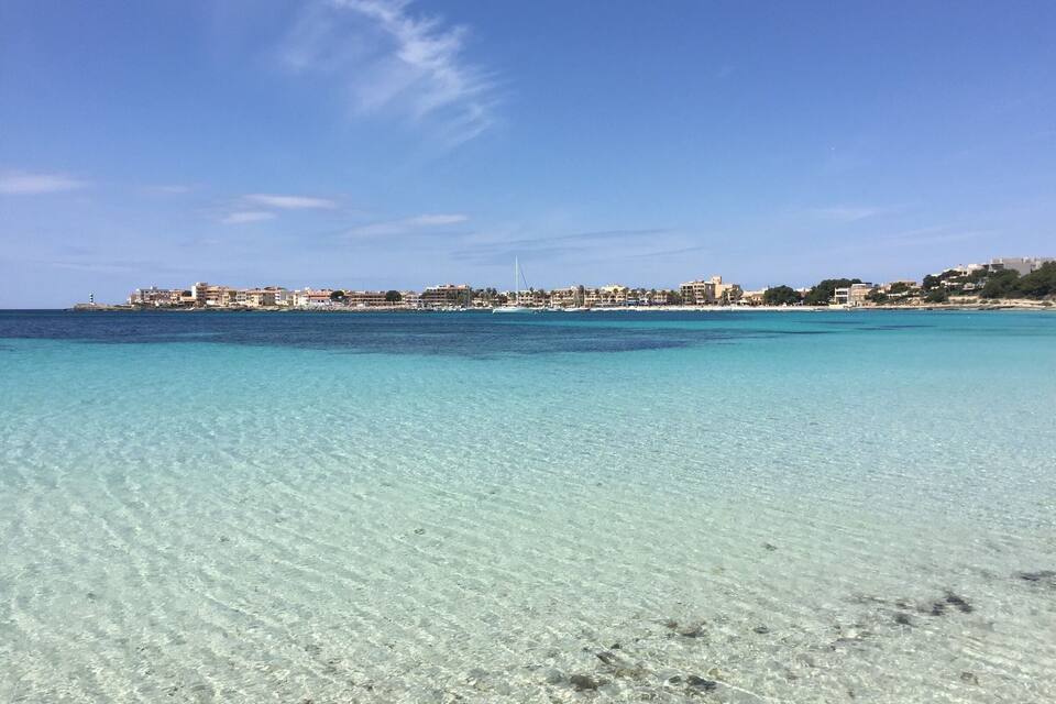 A clear turquoise sea with a sandy beach and a coastal town in the background.