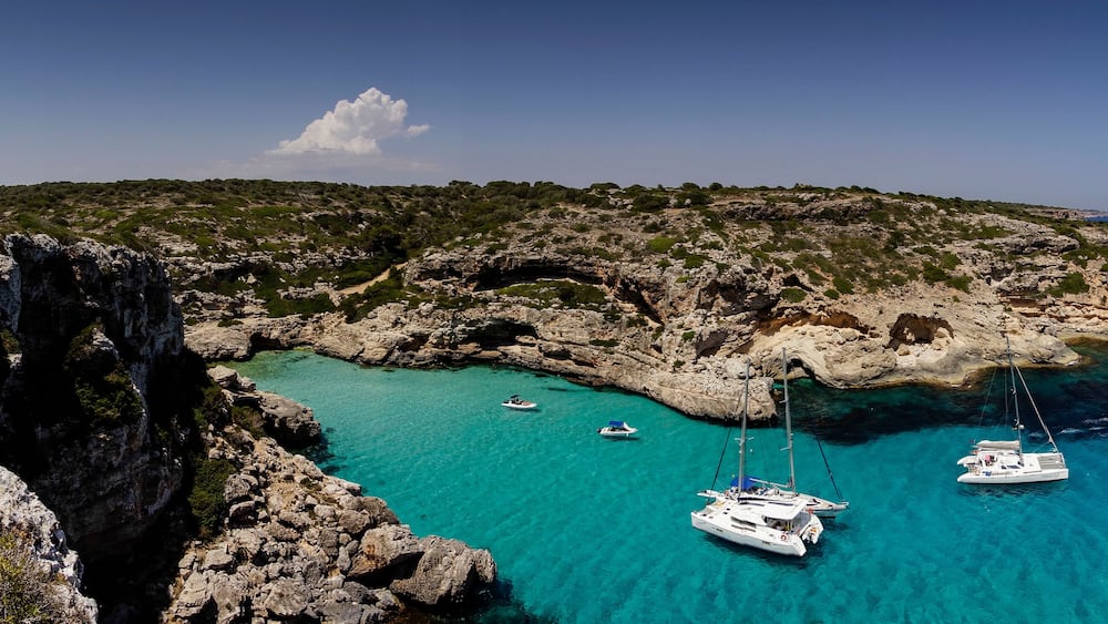 A coastal landscape with rocky cliffs, clear turquoise waters, and several anchored boats.