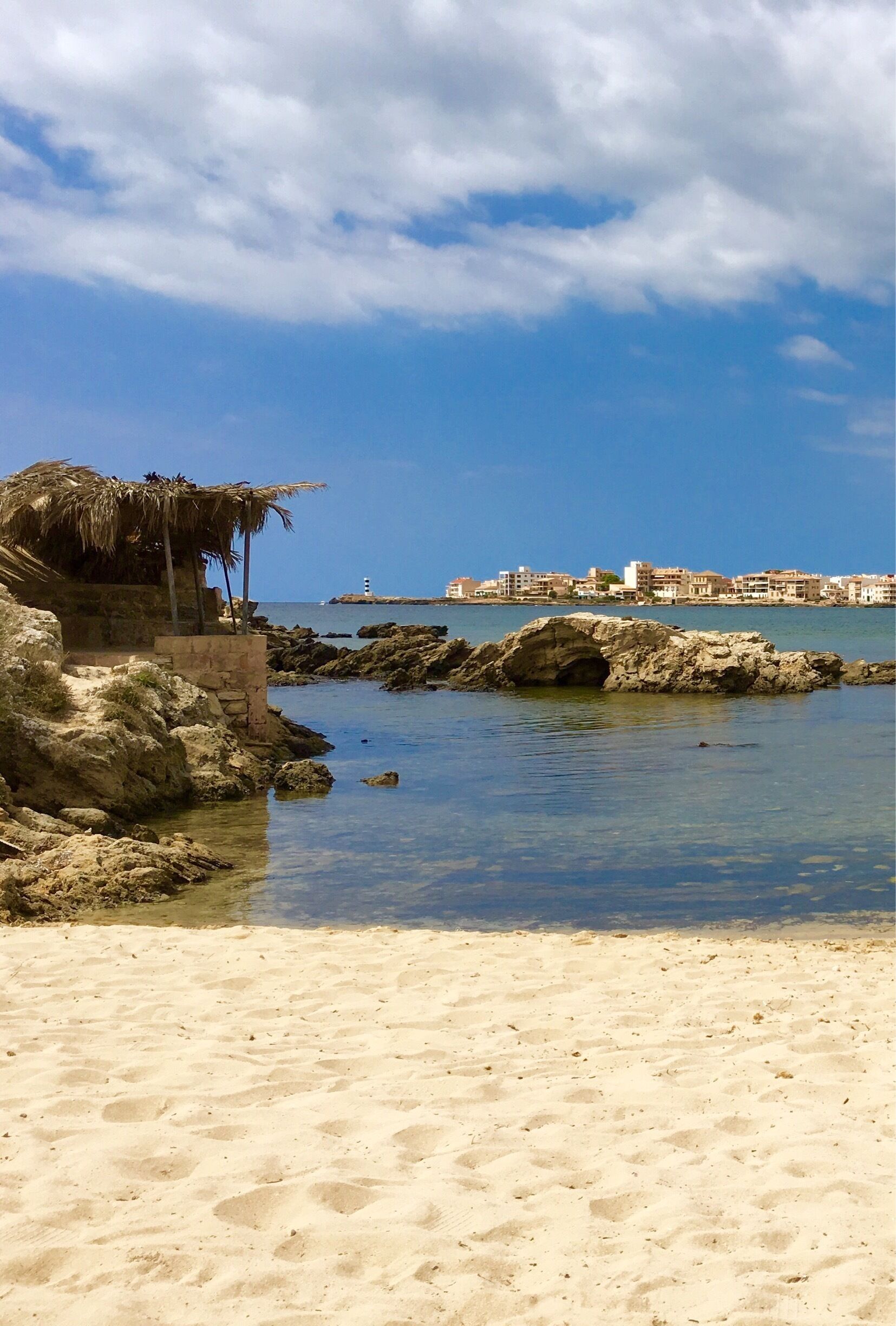 A beach with clear water, a rocky outcrop, and a distant cityscape.