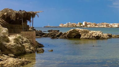 Una playa con agua cristalina, un promontorio rocoso y un horizonte urbano a lo lejos.