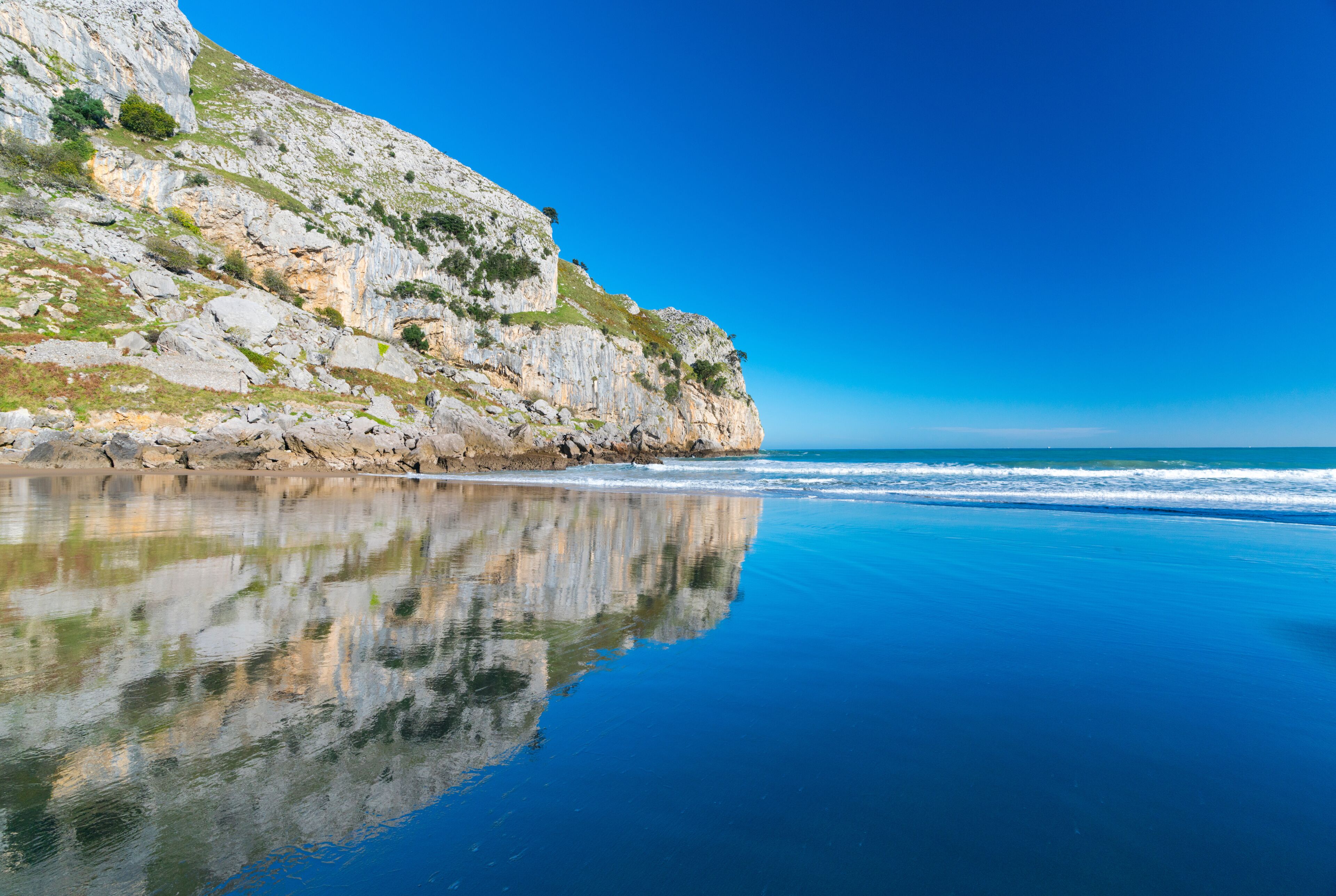 A coastal landscape with a rocky cliff, calm sea, and clear blue sky.