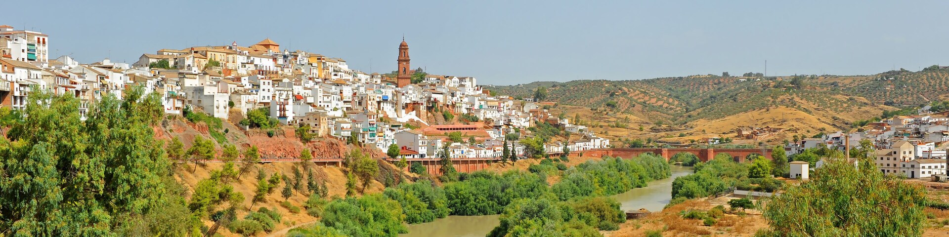 Un pueblo con un río, colinas verdes y una torre destacada.