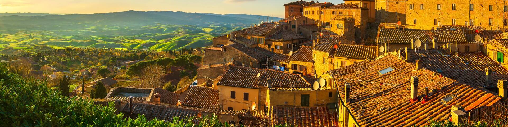 A historic town with terracotta-roofed buildings, a prominent church with a dome, and rolling green hills in the background.