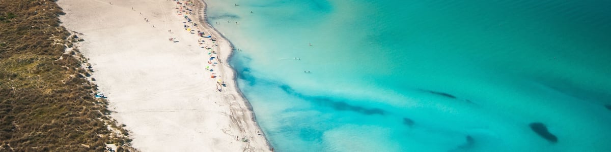An aerial view of a beach with clear turquoise waters, a sandy shore, and a bridge in the background.