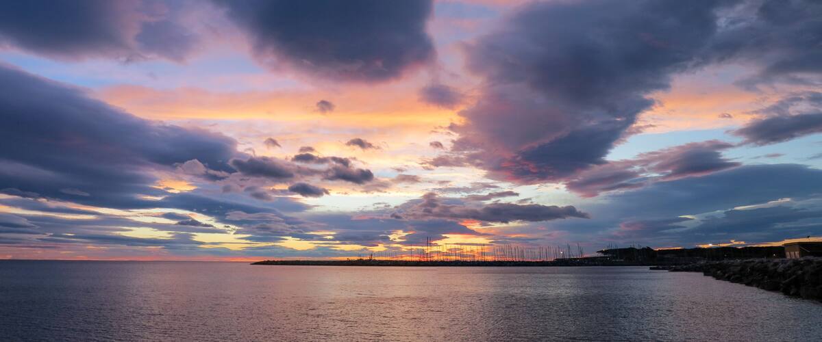 A sunset over a calm sea with a pier extending into the water, surrounded by a rocky coastline.