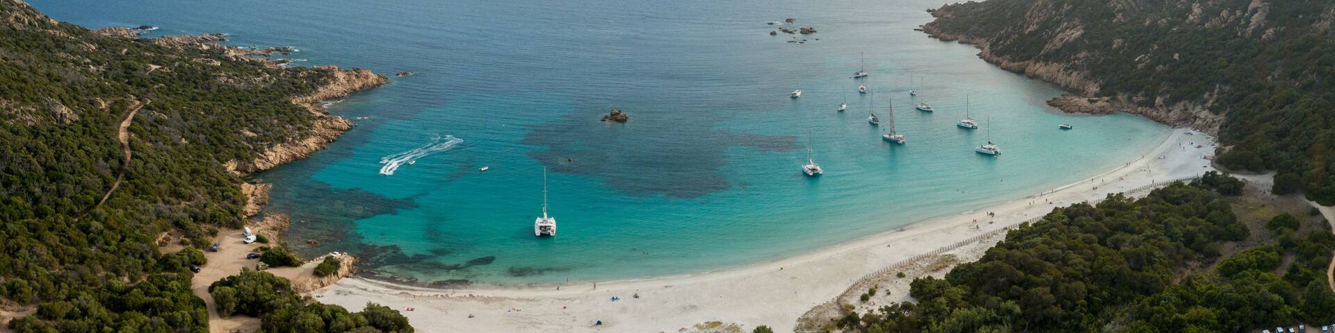 A coastal landscape with a sandy beach, clear turquoise waters, and several boats anchored near the shore.
