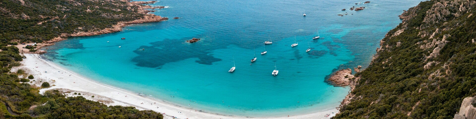 A coastal landscape with a sandy beach, clear turquoise waters, and several boats anchored near the shore.