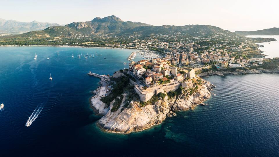 An aerial view of a coastal town with a fortified island, clear blue waters, and mountainous terrain in the background.
