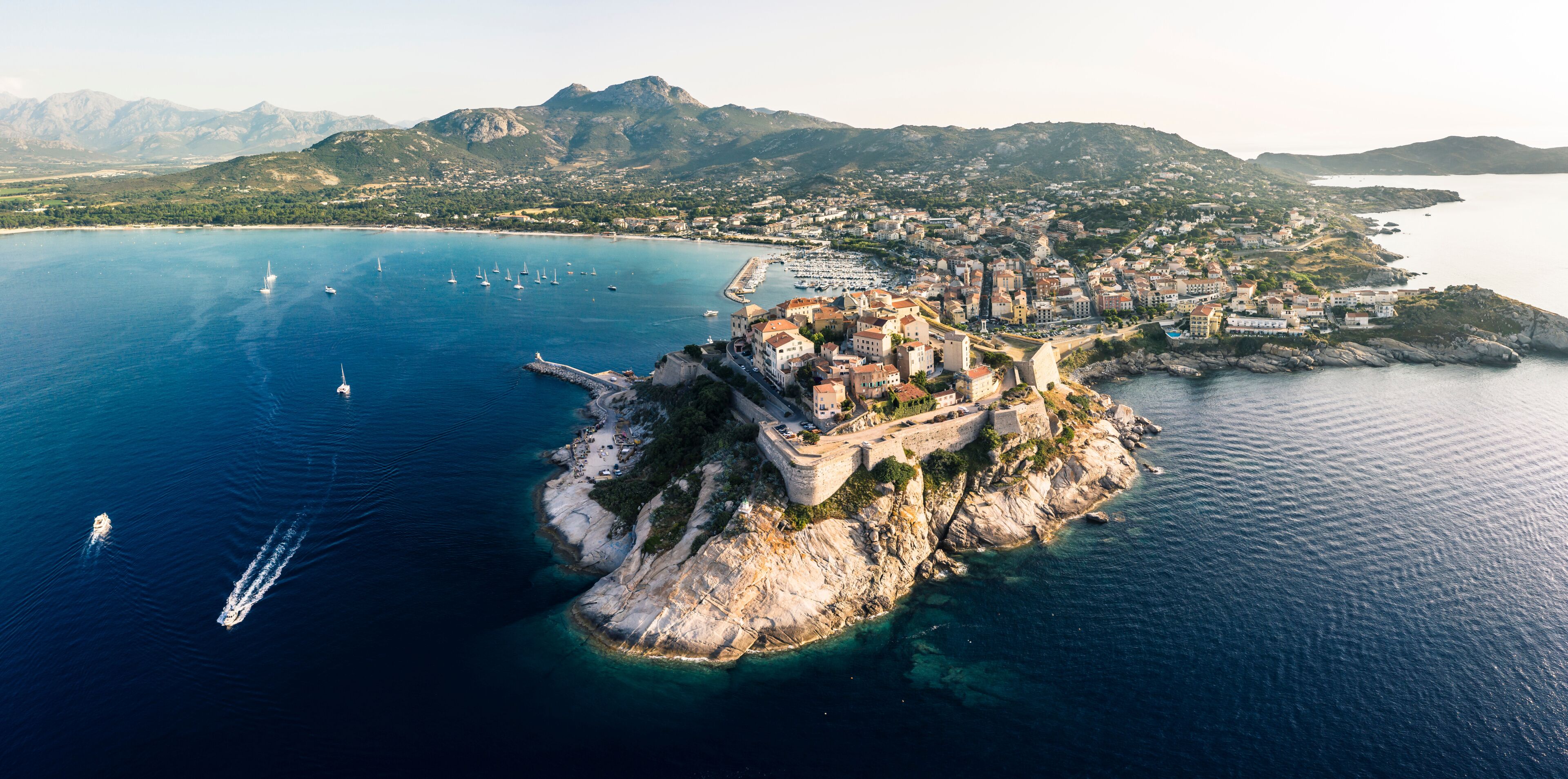 An aerial view of a coastal town with a fortified island, clear blue waters, and mountainous terrain in the background.