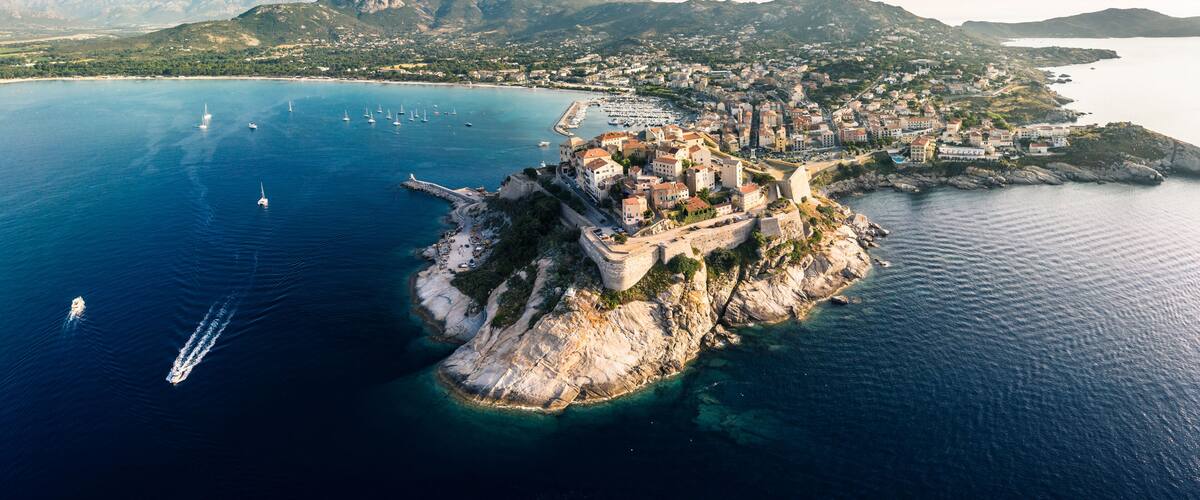 An aerial view of a coastal town with a fortified island, clear blue waters, and mountainous terrain in the background.