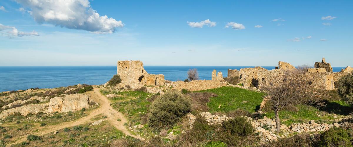 Ancient stone ruins, a clear blue sky, and a distant view of the sea.