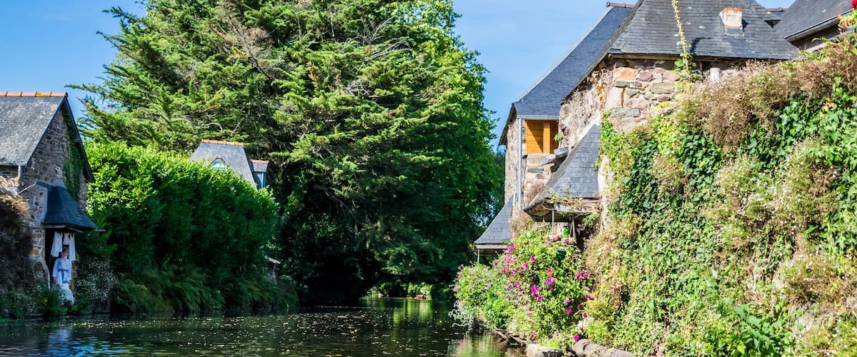 A narrow waterway flanked by stone buildings with steep roofs, surrounded by lush greenery and a large tree.