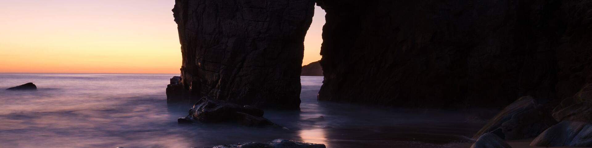 A coastal landscape with a prominent rock formation resembling an arch, smooth sand, and scattered boulders.
