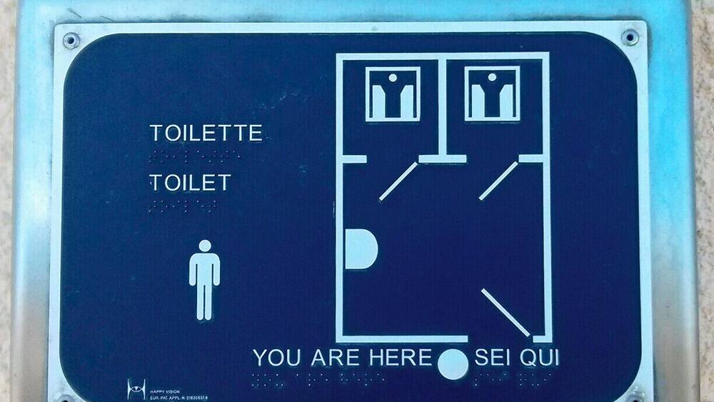 A restroom sign with a male figure, a toilet symbol, and text in multiple languages.