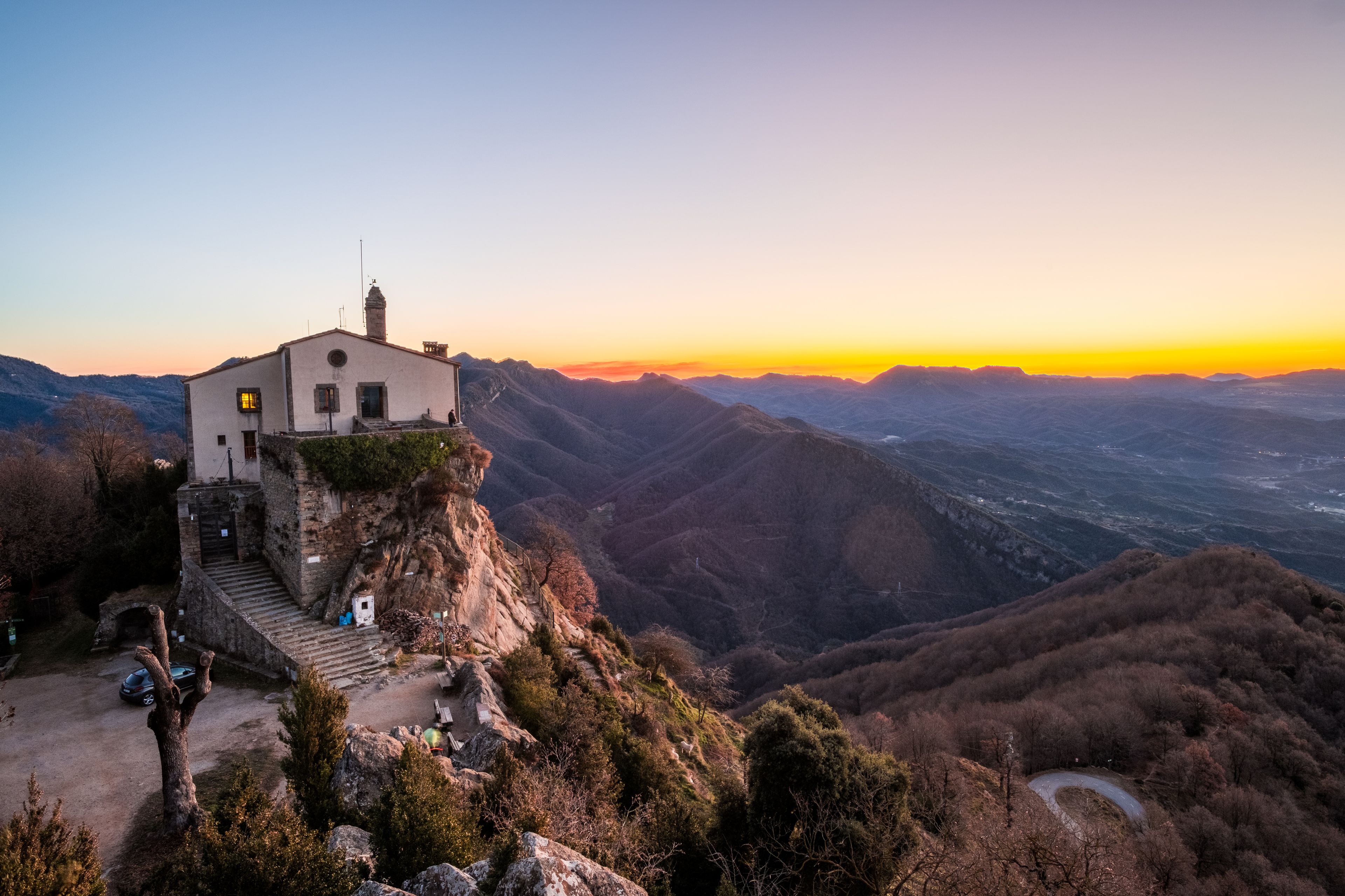 Un edificio blanco con chimenea, ubicado en una ladera con vistas a un valle y montañas al fondo.