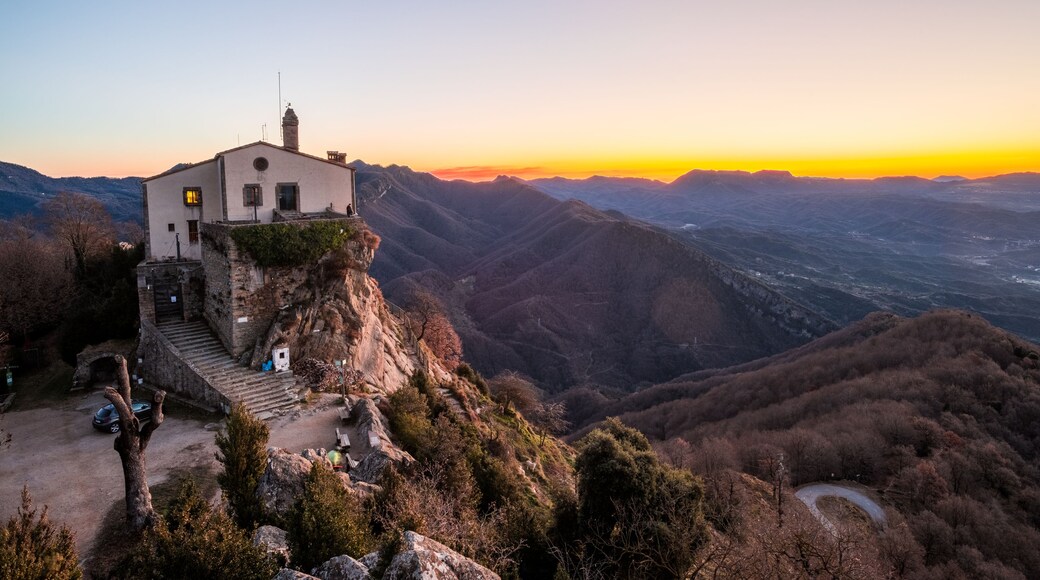 Un edificio blanco con chimenea, ubicado en una ladera con vistas a un valle y montañas al fondo.