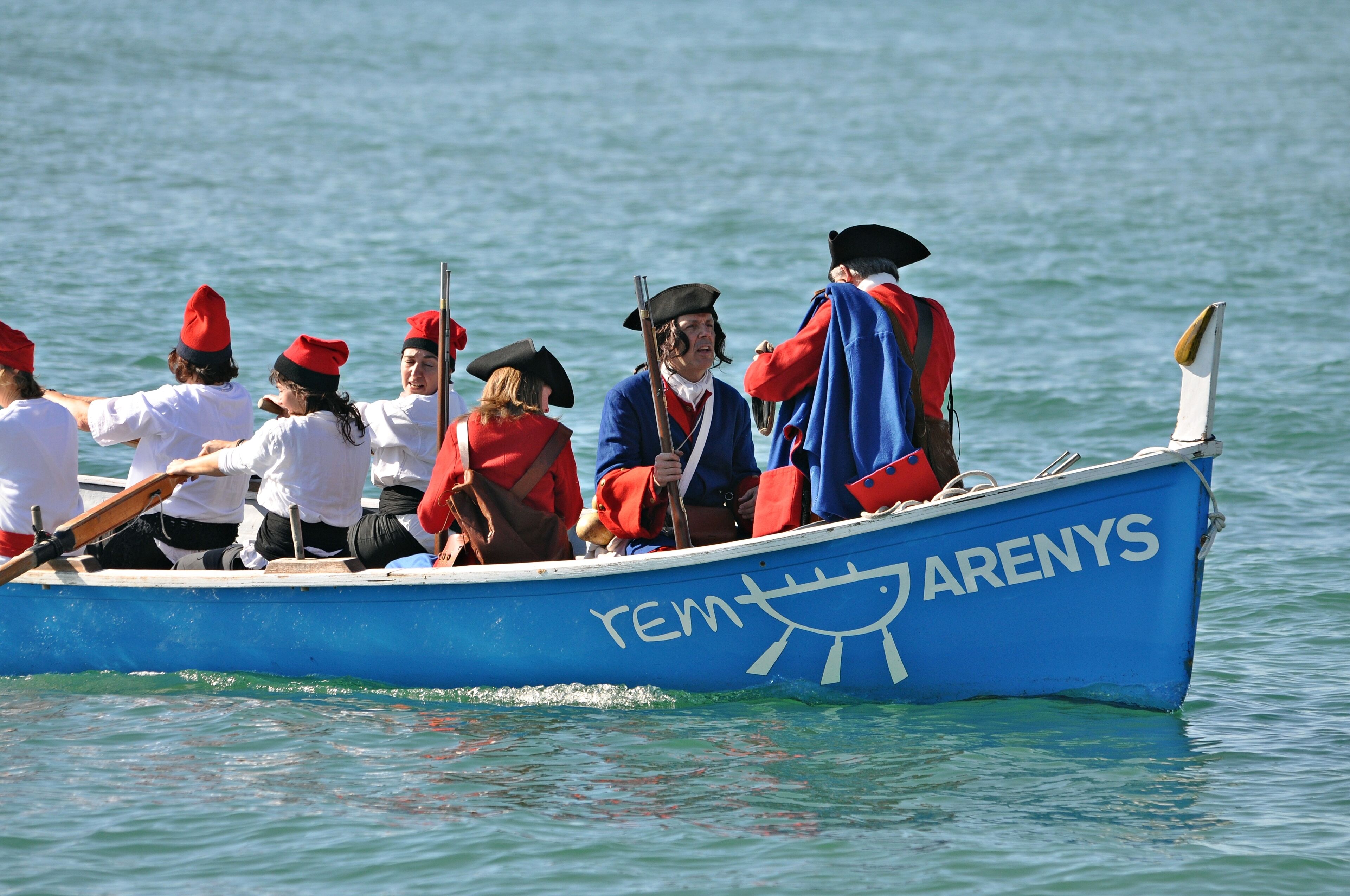 A blue boat with 'rem' and 'arenys' written on it, with people rowing and wearing red hats.
