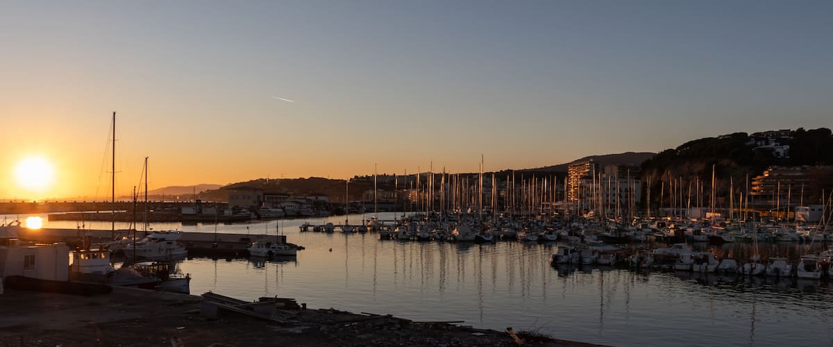 A marina at sunset with numerous boats docked, a clear sky, and buildings in the background.
