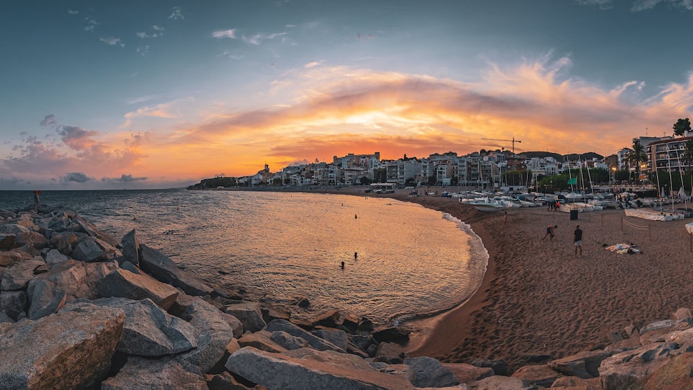 A beach with rocky shoreline, boats, and buildings in the background during sunset.